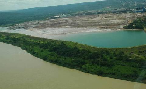 Fotografia da Barragem de rejeitos minerários da Votorantim Metais, em Três Marias, ao lado do rio São Francisco. Ao fundo, barragem, cor verde. Embaixo, água suja do rio São Francisco. Um técnico em mineração me disse: “Construir uma barragem de rejeitos de mineração com metais pesados na beira de um rio é um absurdo, um crime de lesa-pátria.” Isso a Votorantim fez, após jogar os rejeitos minerários diretamente no rio São Francisco durante 14 anos.